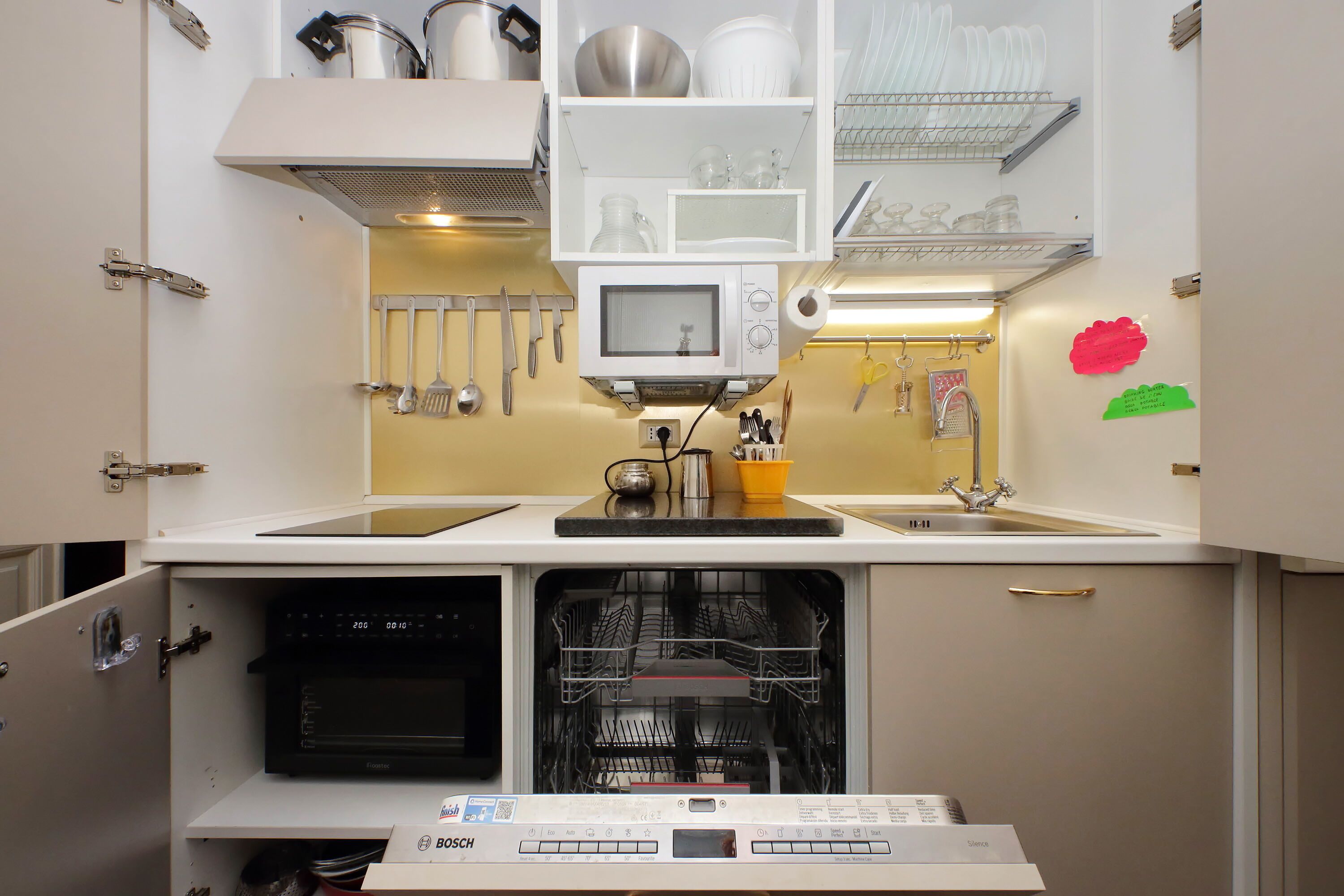 Cocina compacta con lavavajillas Bosch, microondas y varios utensilios colgados en la pared.