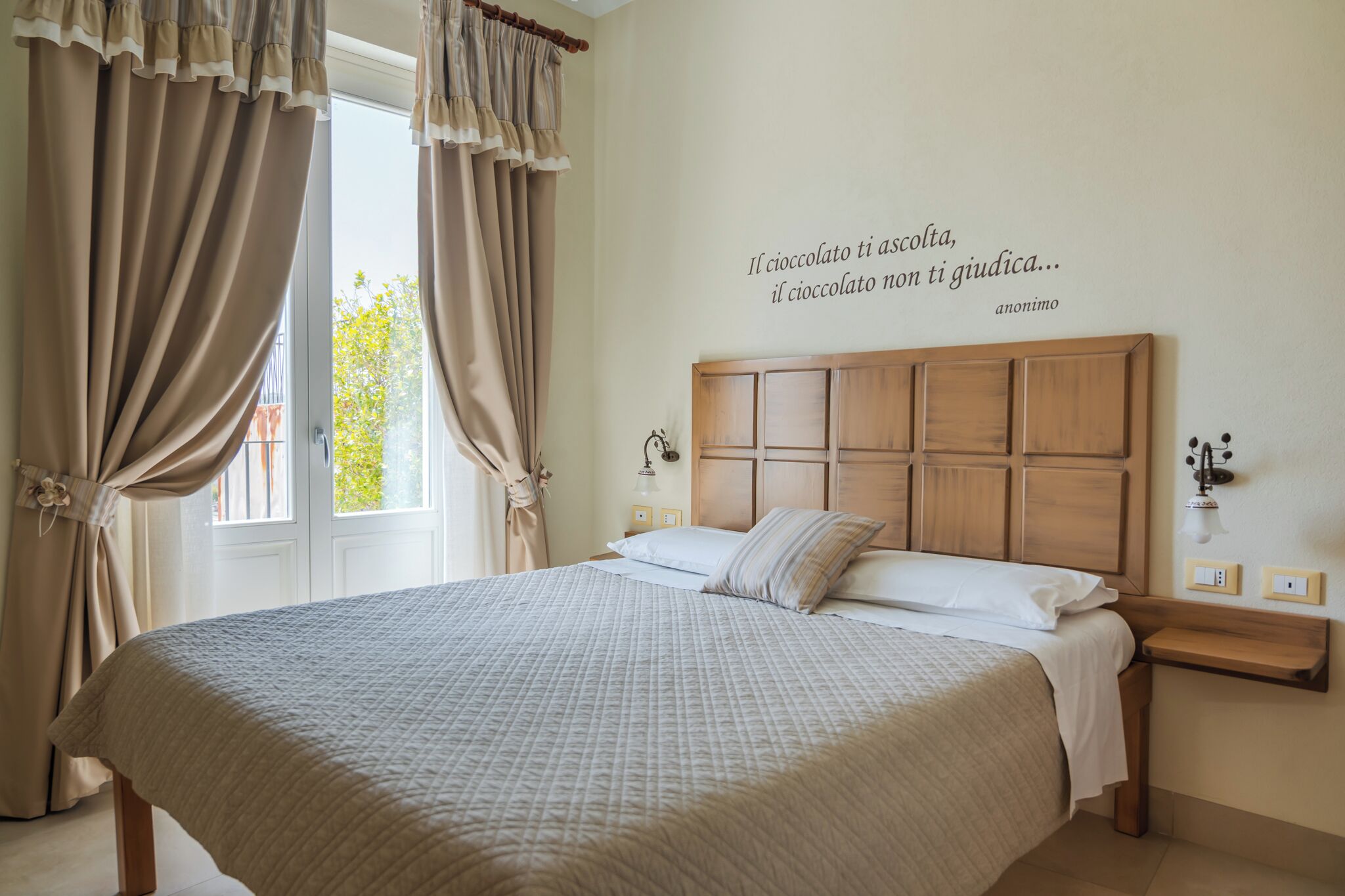 Un dormitorio con una cama grande, cortinas, cabecera, lámparas de noche y una frase decorativa en la pared.