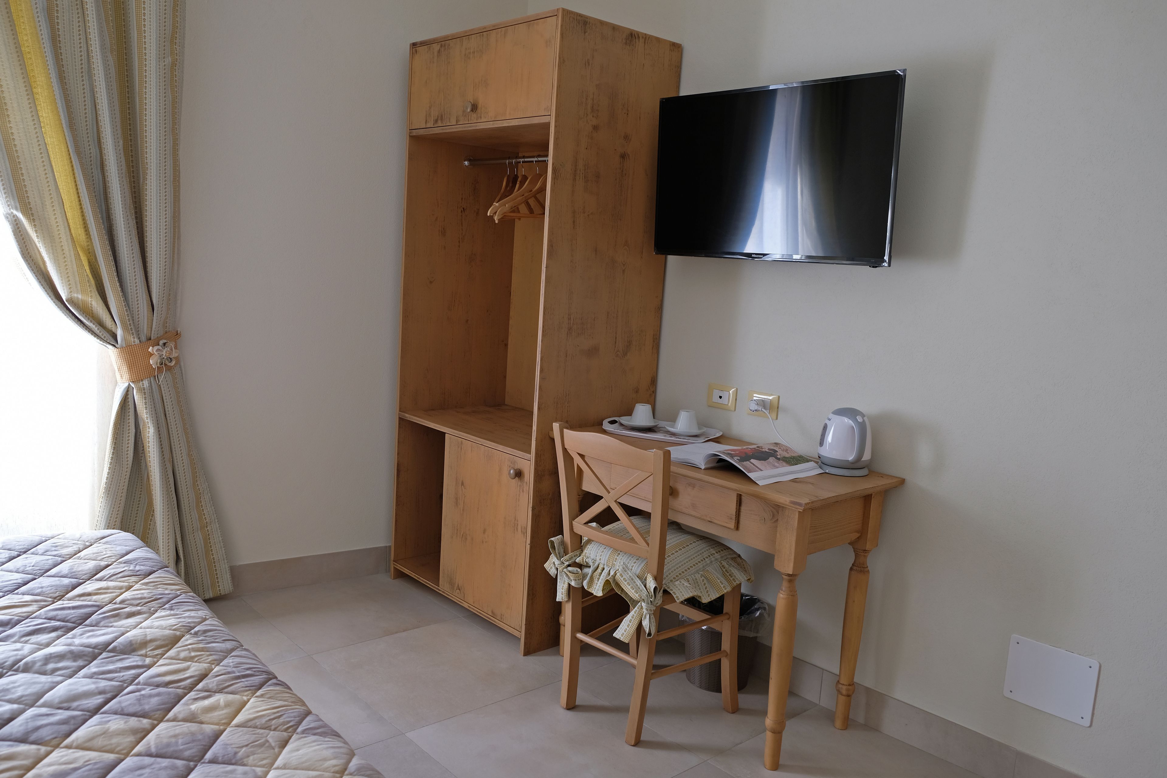 A bedroom with a bed, a wooden wardrobe, a desk with a chair, a television, and a window with curtains.