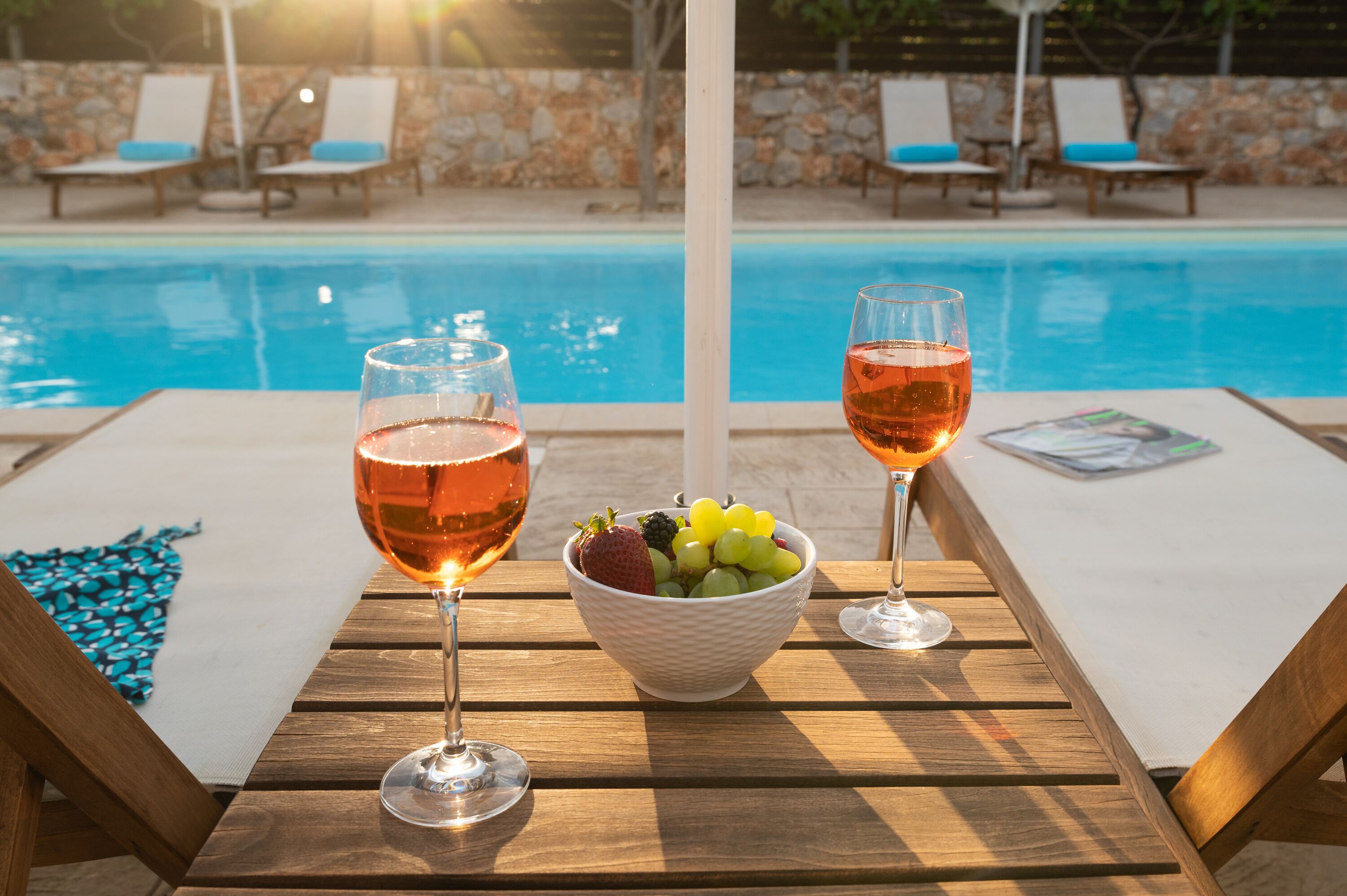 Una mesa junto a la piscina con dos copas de vino rosado, un bol de fruta y una revista.