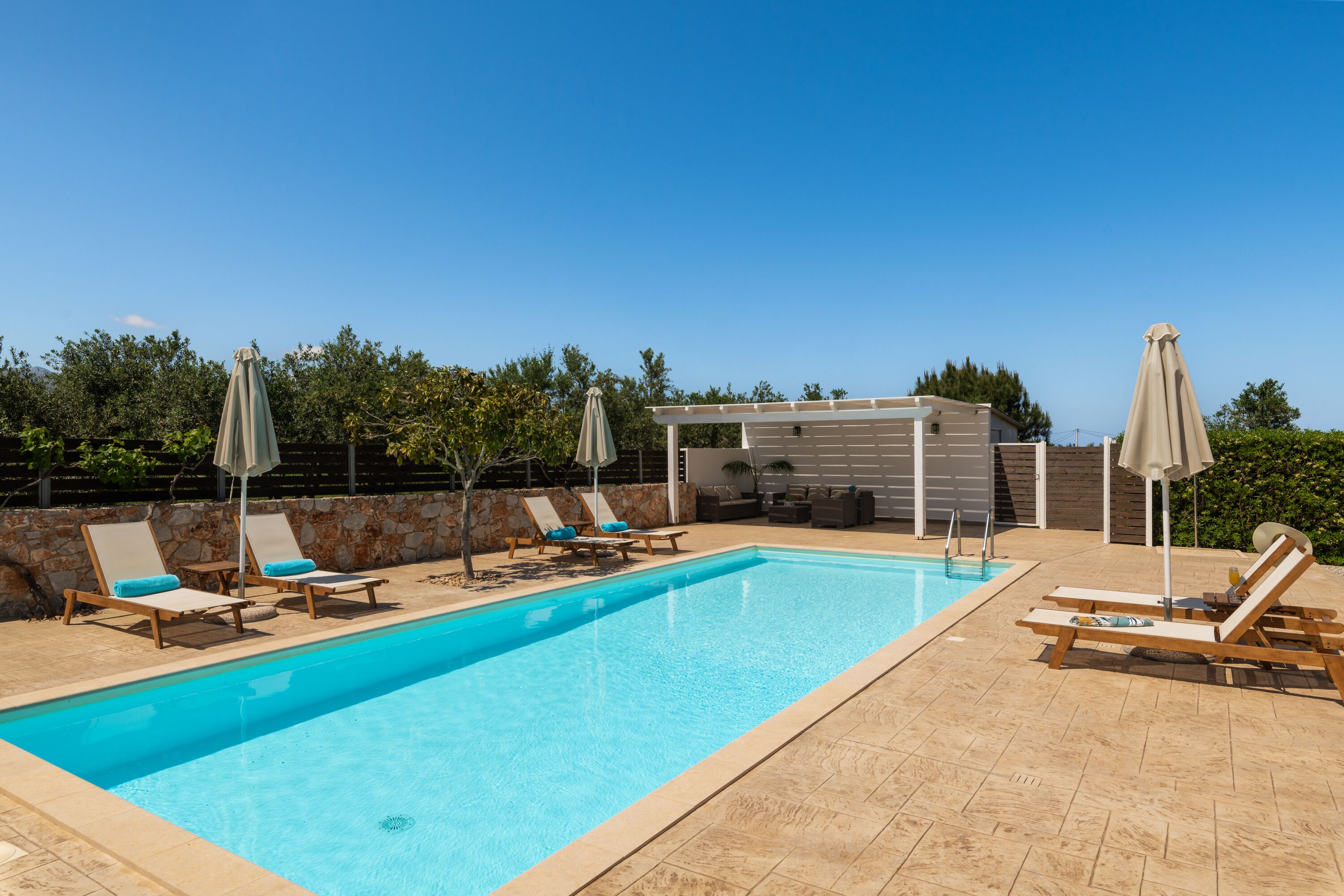 Una piscina rectangular con tumbonas y sombrillas, rodeada de un muro de piedra y follaje.