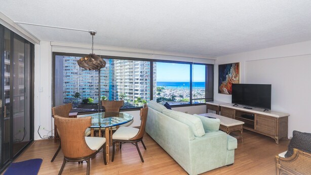 A modern living room with a large window, a sofa, a dining table, and a TV. - Perfect Waikiki Location, Right Near The Beach! (Honolulu)
