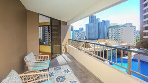 A balcony with wicker furniture, a glass door, and a view of a cityscape and swimming pool. - Perfect Waikiki Location, Right Near The Beach! (Honolulu)