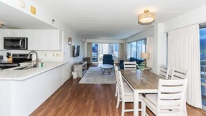Une cuisine moderne équipée d’un four à micro-ondes, d’un évier et de meubles blancs, qui donne sur un coin repas avec une table en bois et des chaises blanches, et offre une vue sur de hauts immeubles à travers de grandes fenêtres.