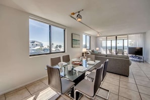 A modern living room with a glass dining table, grey sofas, and a large TV.