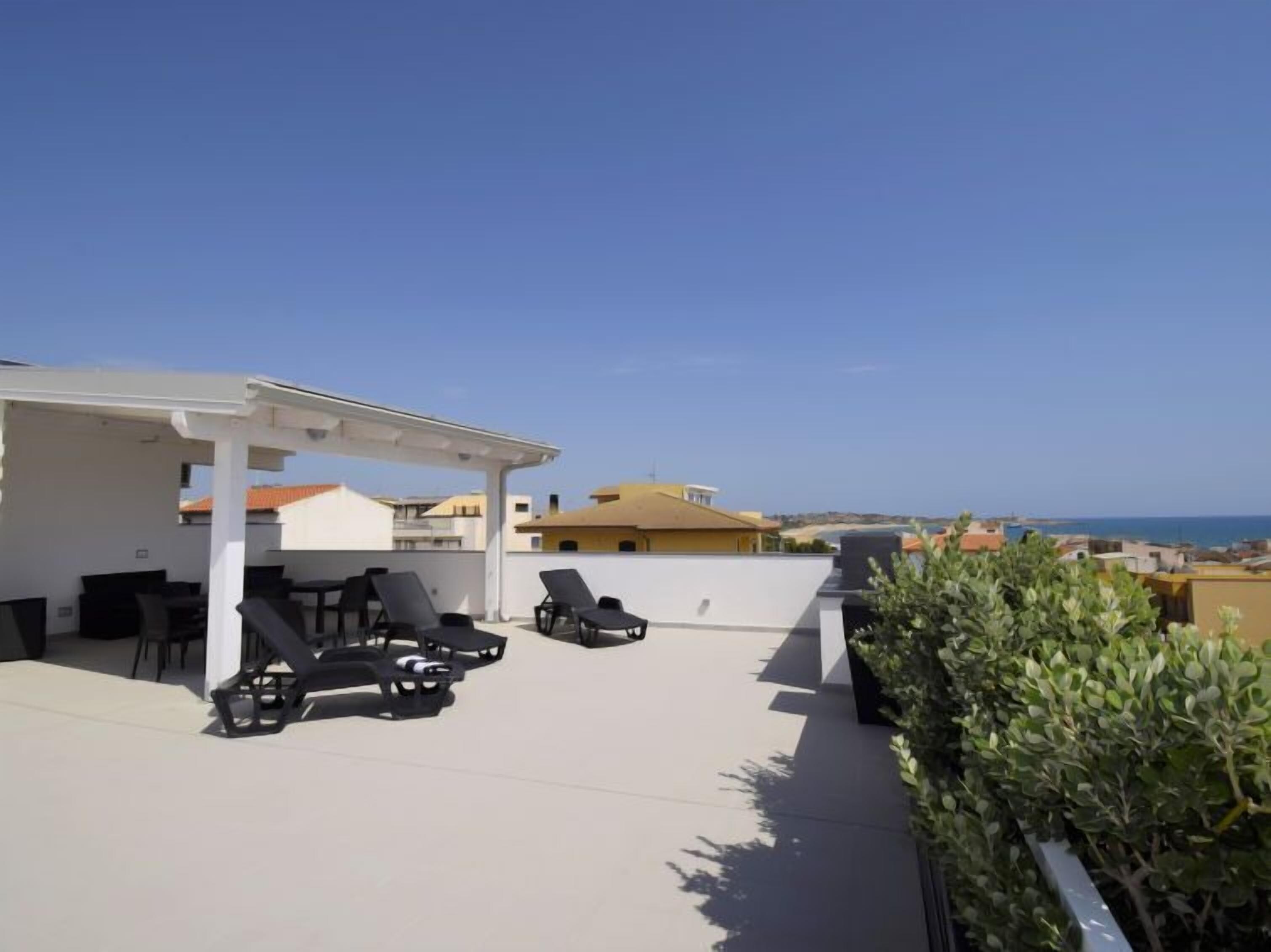 A rooftop terrace with lounge chairs and a covered seating area, overlooking a coastal cityscape.