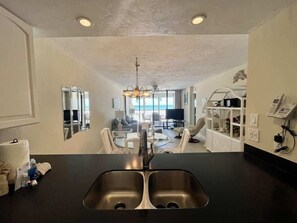 A modern kitchen with a double sink, a dining area with a round table and chairs, and a view of the ocean through large windows.