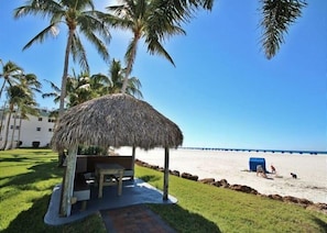 A beachside hut with a thatched roof, a table, and chairs, surrounded by palm trees and a clear view of the ocean.