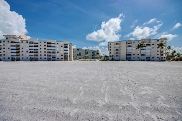 Un hotel frente a la playa con varios pisos, una playa de arena en primer plano y palmeras.