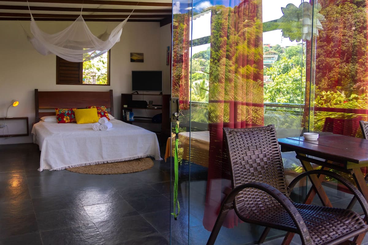 Une chambre à coucher avec un lit, une table de chevet et un téléviseur. Il y a une fenêtre avec des rideaux et une vue sur la verdure à l’extérieur.