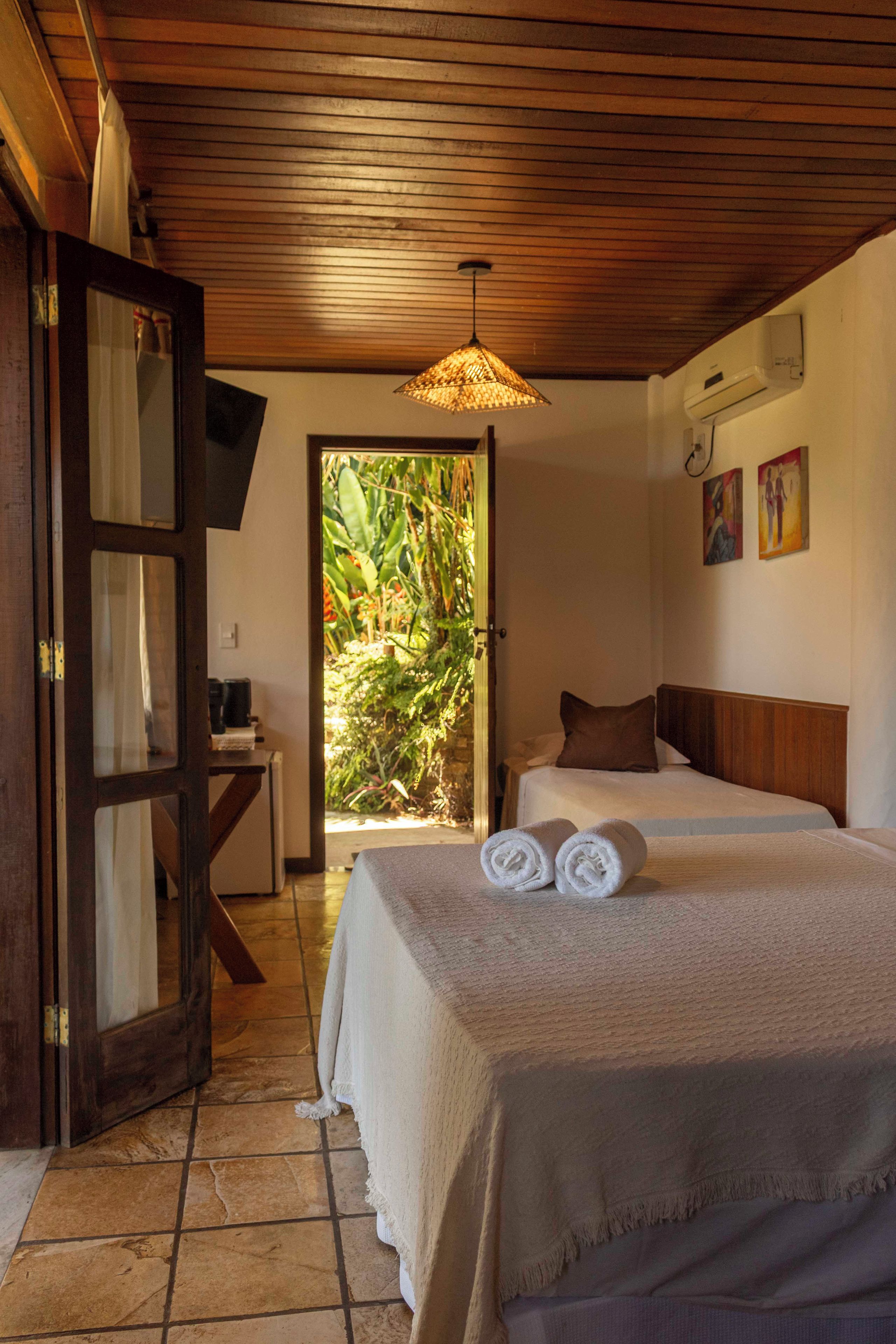 A room with two beds, a wooden ceiling, a door with a mirror, and a view of greenery outside.