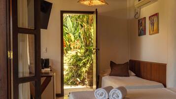 A room with two beds, a wooden ceiling, a door with a mirror, and a view of greenery outside.