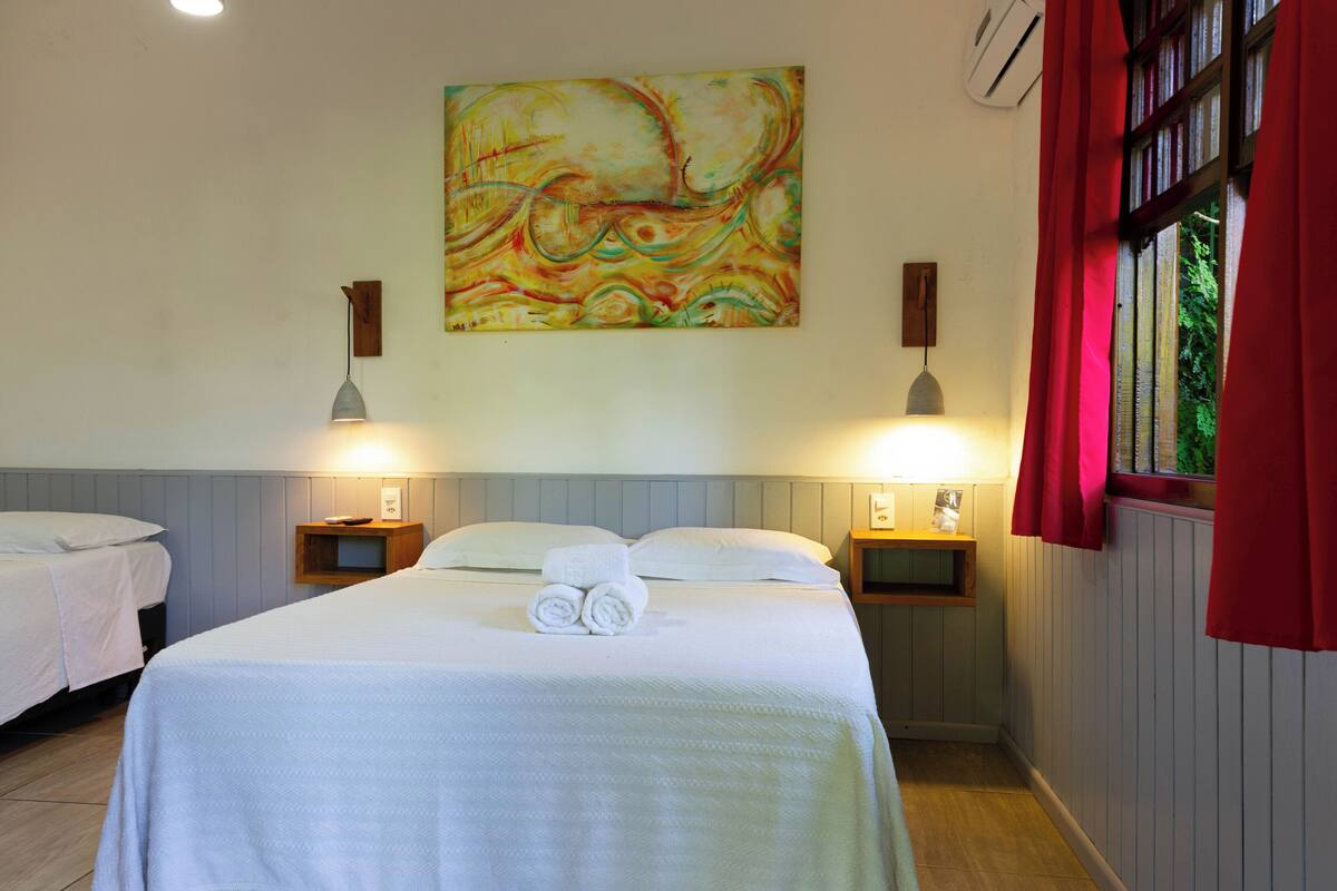 Une chambre d’hôtel avec deux lits, un tableau au mur et une fenêtre avec des rideaux rouges.