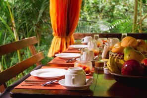 Une table de salle à manger en bois, mise avec des assiettes, des couverts, un saladier rempli de fruits et une tasse, le tout entouré d’une végétation luxuriante.