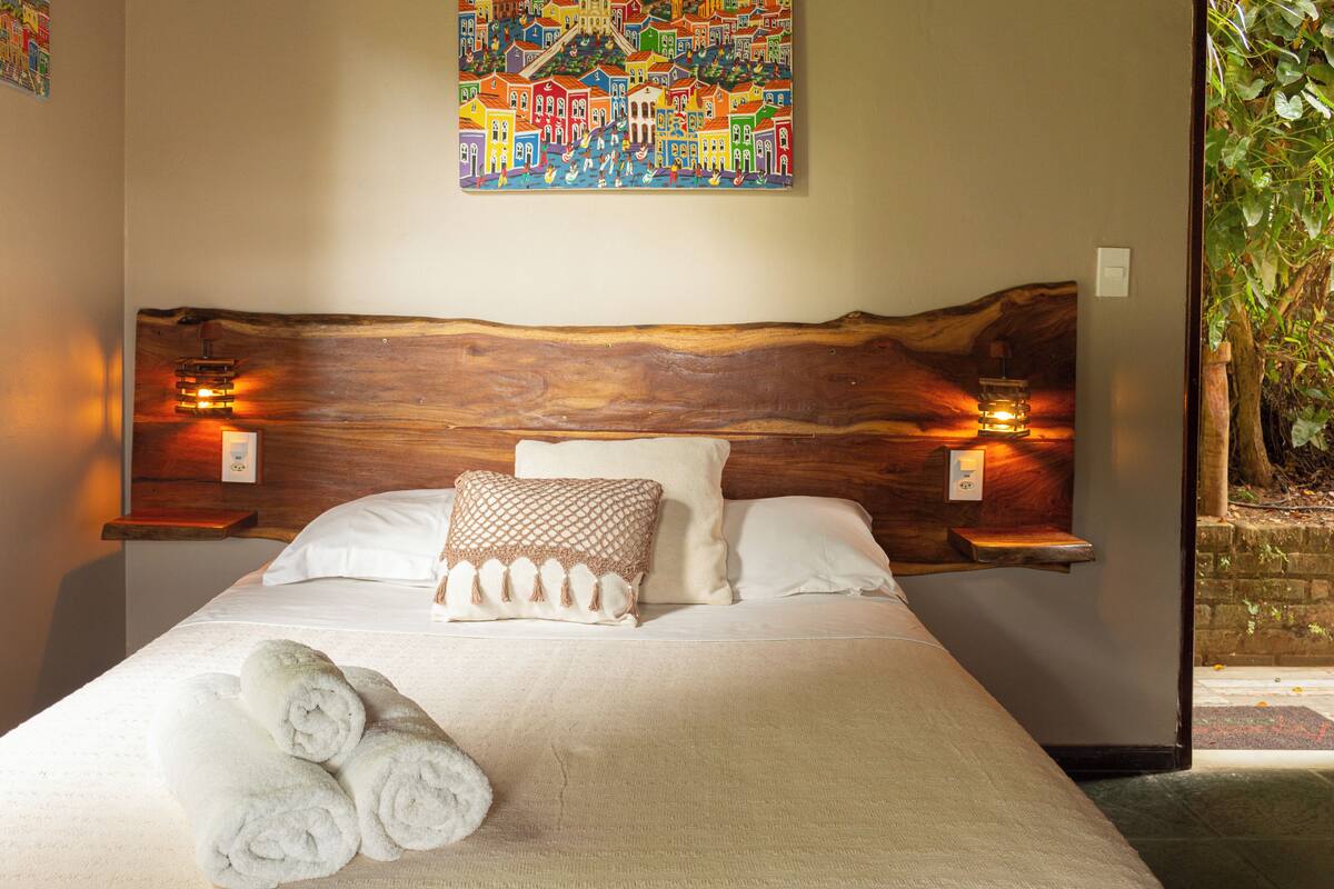 Un lit bien fait, avec du linge de lit blanc et des serviettes pliées, une tête de lit en bois agrémentée de lampes fixées au mur, et un tableau coloré au-dessus.