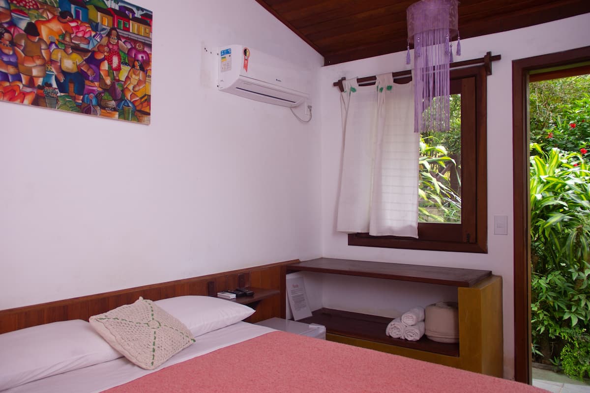 Une chambre à coucher avec un lit, une table de chevet, une étagère avec des serviettes, une fenêtre avec des rideaux, un climatiseur et un tableau coloré accroché au mur.