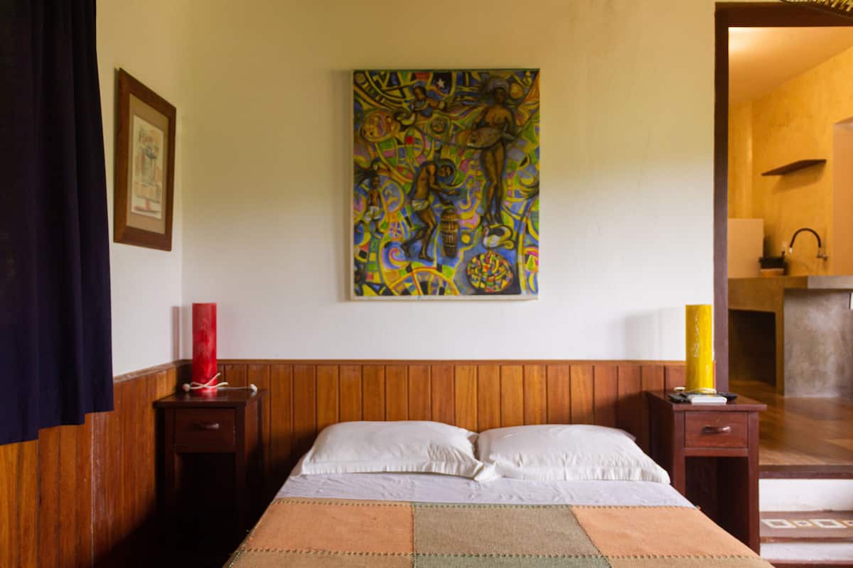 Une chambre d’hôtel avec une tête de lit en bois, un lit recouvert de draps blancs, une peinture abstraite aux couleurs vives accrochée au mur, et une table de chevet ornée d’un vase rouge et d’une lampe jaune.