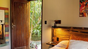 Une chambre avec une porte en bois, un lit recouvert de draps blancs, une table de chevet avec une lampe, et une vue sur la verdure à l’extérieur.