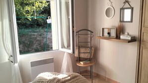 A bedroom with a bed, a chair, a shelf with decorative items, and a window with curtains. - PRETTY PROVENCAL VILLA IN THE HEART OF THE LUBÉRON PARK 7 MIN FROM GORDES (Saint-Saturnin-lès-Apt)