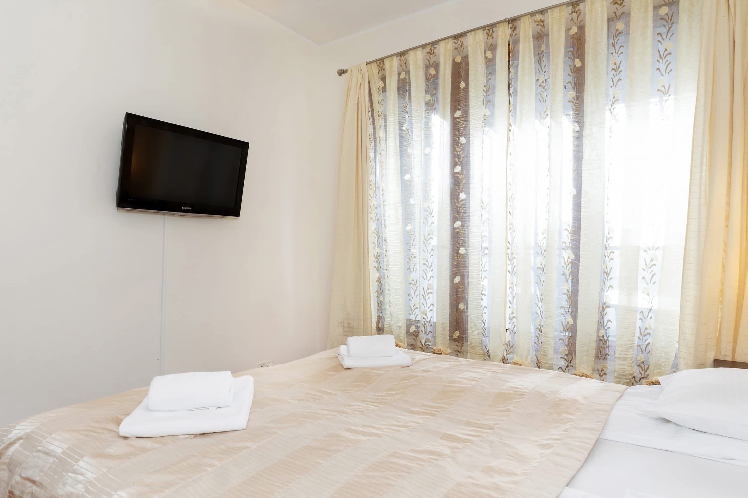 A hotel room with a bed, a flat-screen TV mounted on the wall, and a window with floral curtains.