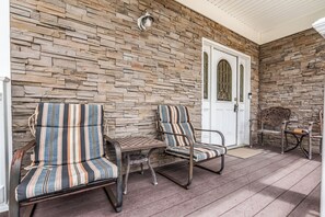 A covered porch with two striped chairs and a small table, a stone wall, and a white door with a glass window.