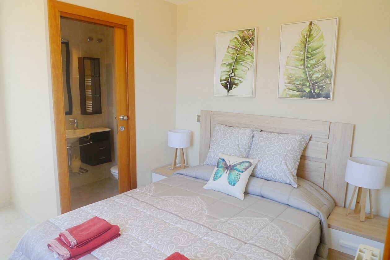 Een slaapkamer met een bed, twee wandlampen, een nachtkastje, een spiegel en twee ingelijste schilderijen van bladeren.