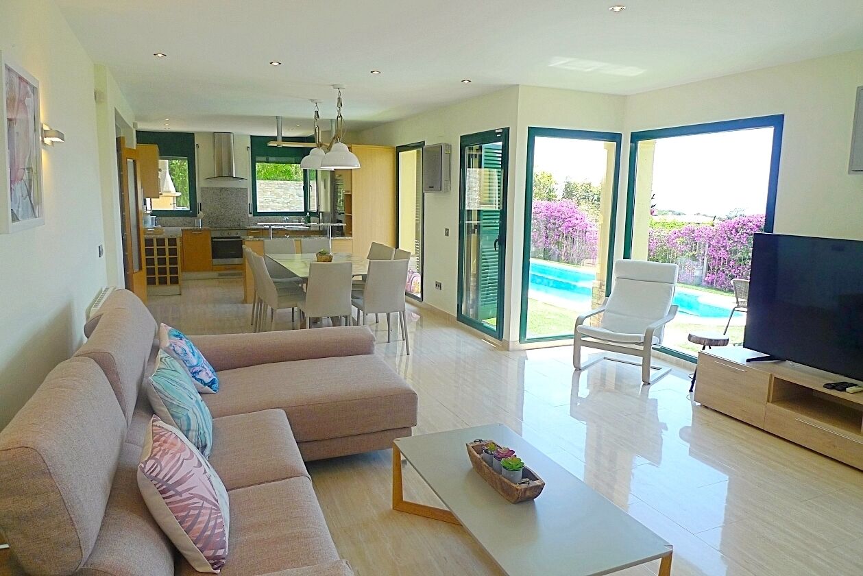 Un salon moderne avec un canapé, une table basse et un téléviseur. Il y a également un coin repas avec des chaises et une table. La pièce est dotée de grandes fenêtres et offre une vue sur une piscine.