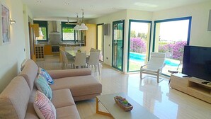 A modern living room with a sofa, coffee table, and TV. There is a dining area with chairs and a table. The room has large windows and a view of a swimming pool.
