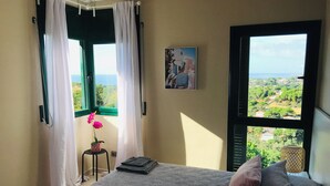 A bedroom with a bed, a window with a view of greenery, a small table with a flower, and a painting on the wall.