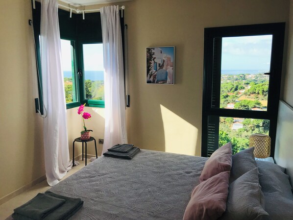 A bedroom with a bed, a window with a view of greenery, a small table with a flower, and a painting on the wall.