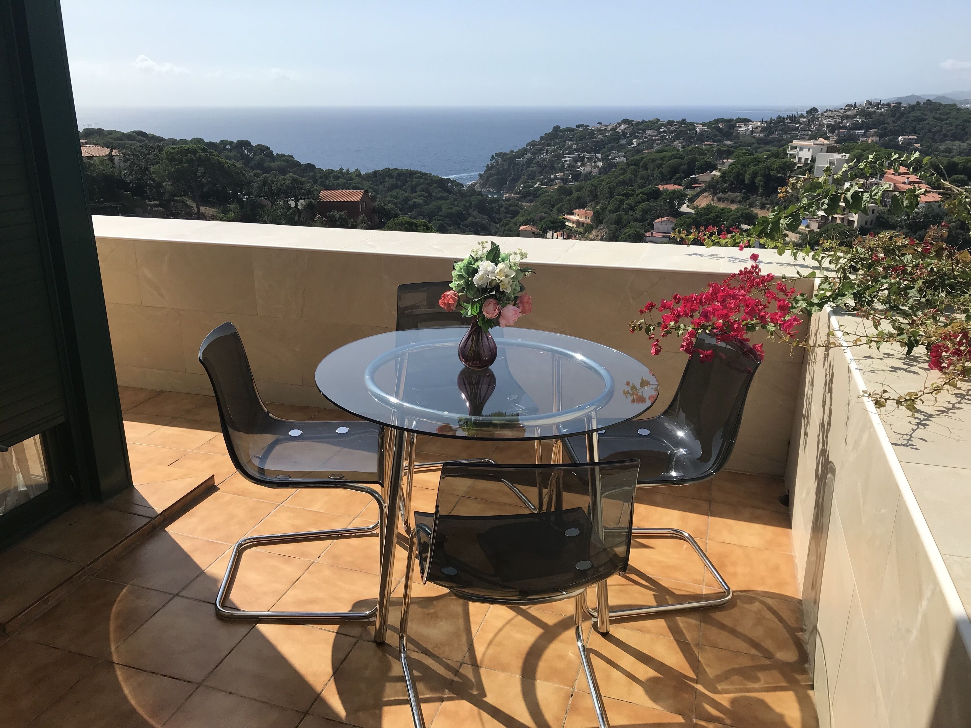 Une table ronde en verre, entourée de chaises en métal, sur un balcon offrant une vue sur la mer et une ville située sur une colline.