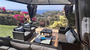 An outdoor patio with wicker furniture, a table, and a view of the sea and hills.