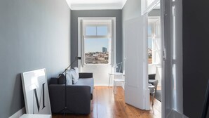 A modern living room with a grey sofa, a small white table, and a view of the city through a large window. - GuestReady - Innvict Admiral (Porto)