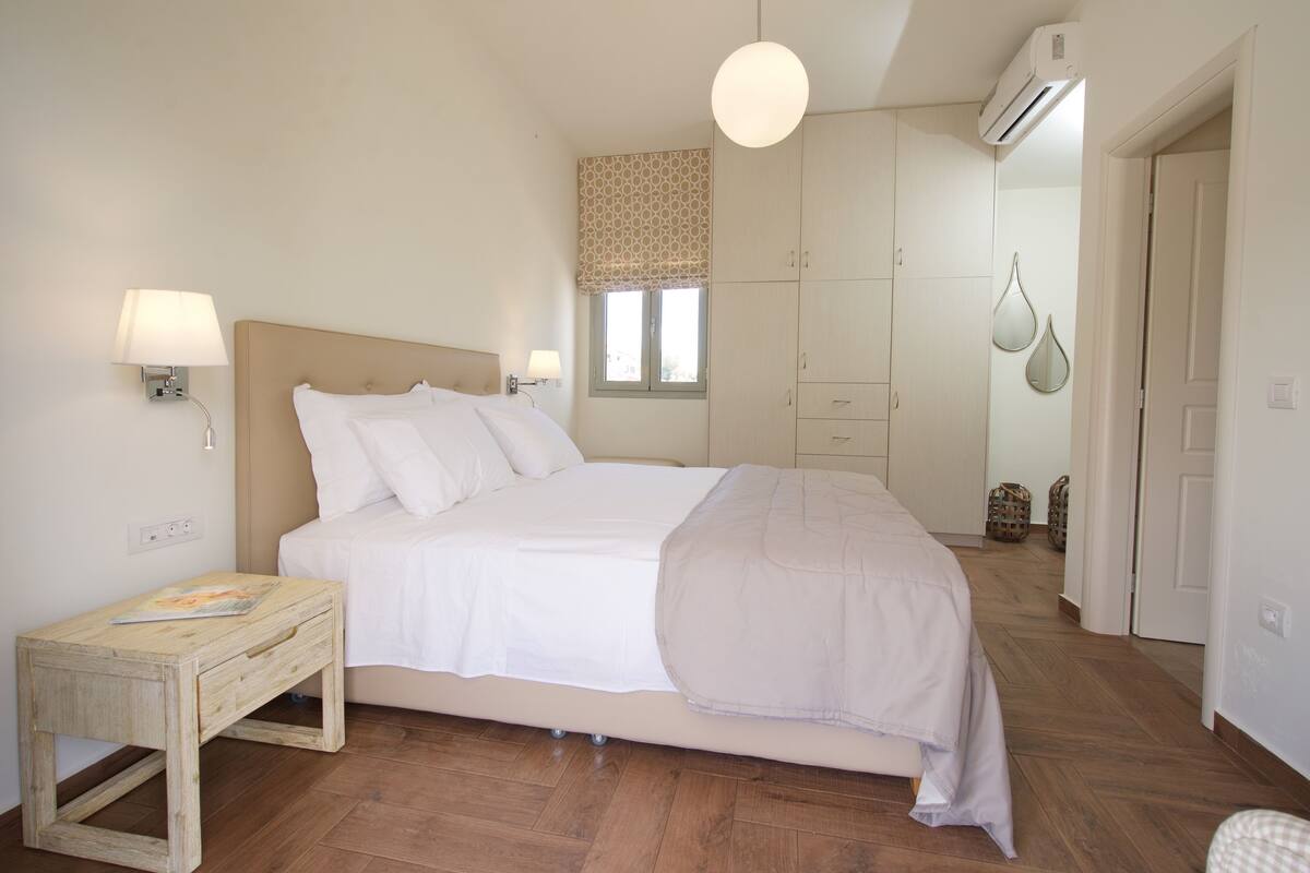 Ein Schlafzimmer mit einem Bett, Nachttischen, einem Fenster mit einem gemusterten Rollo, einer Pendelleuchte und einem Kleiderschrank.