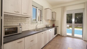 A modern kitchen with light wood cabinets, a microwave, a sink, and a window overlooking a pool. - Amarianos Villas (Kefalonia)