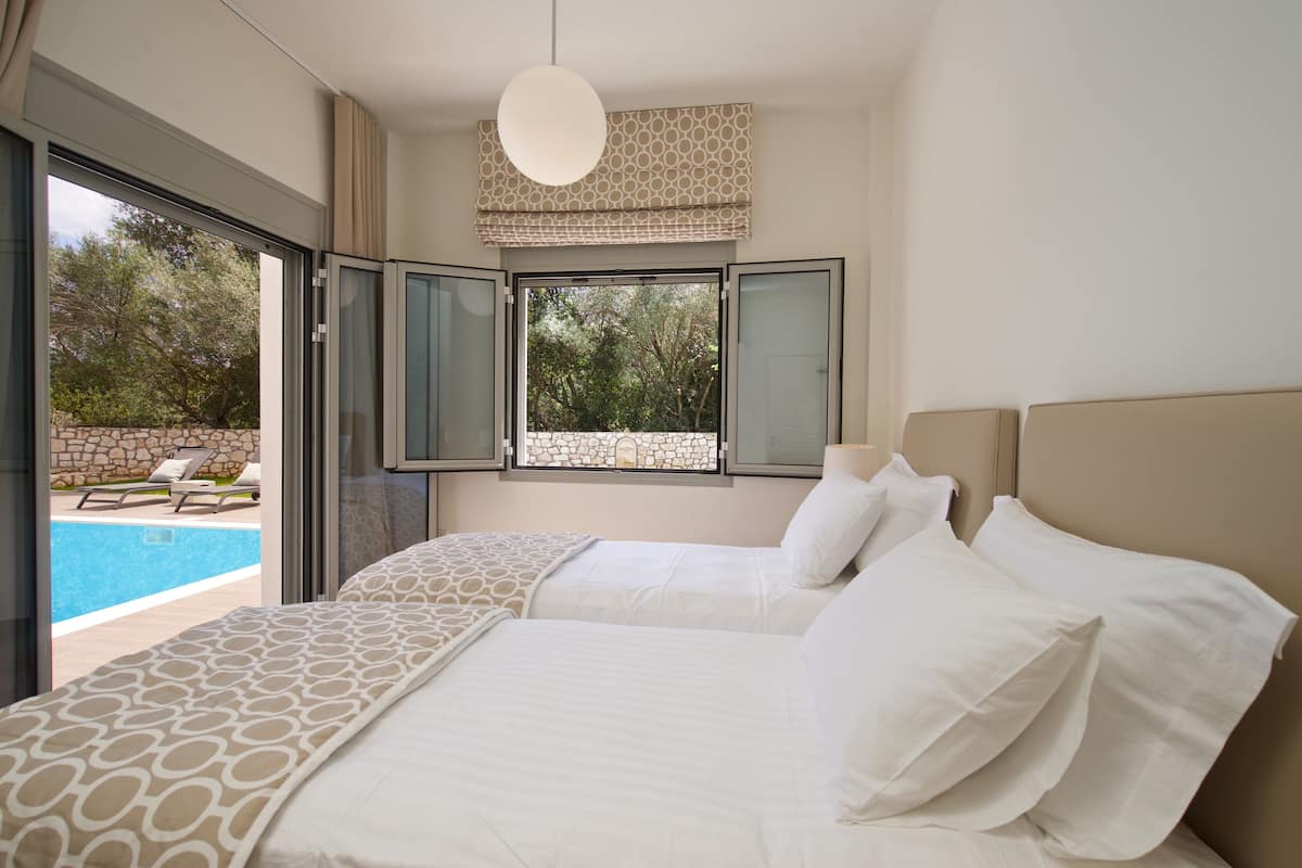 Ein Schlafzimmer mit zwei Betten, einem Fenster mit Blick auf Pool und Bäume sowie einer hängenden Deckenleuchte.