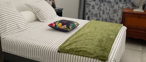 Una cama bien hecha con ropa de cama a rayas, un plaid verde y una almohada colorida.