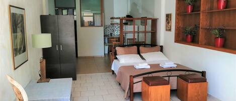 A bedroom with a bed, bedside tables, a desk, a chair, a wardrobe, and a shelf with potted plants.