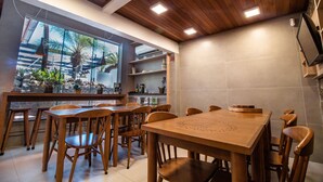 A dining area with wooden tables and chairs, a large window with a view of palm trees, and a ceiling with recessed lighting. - Spazio del Sole Jurere (Florianópolis)