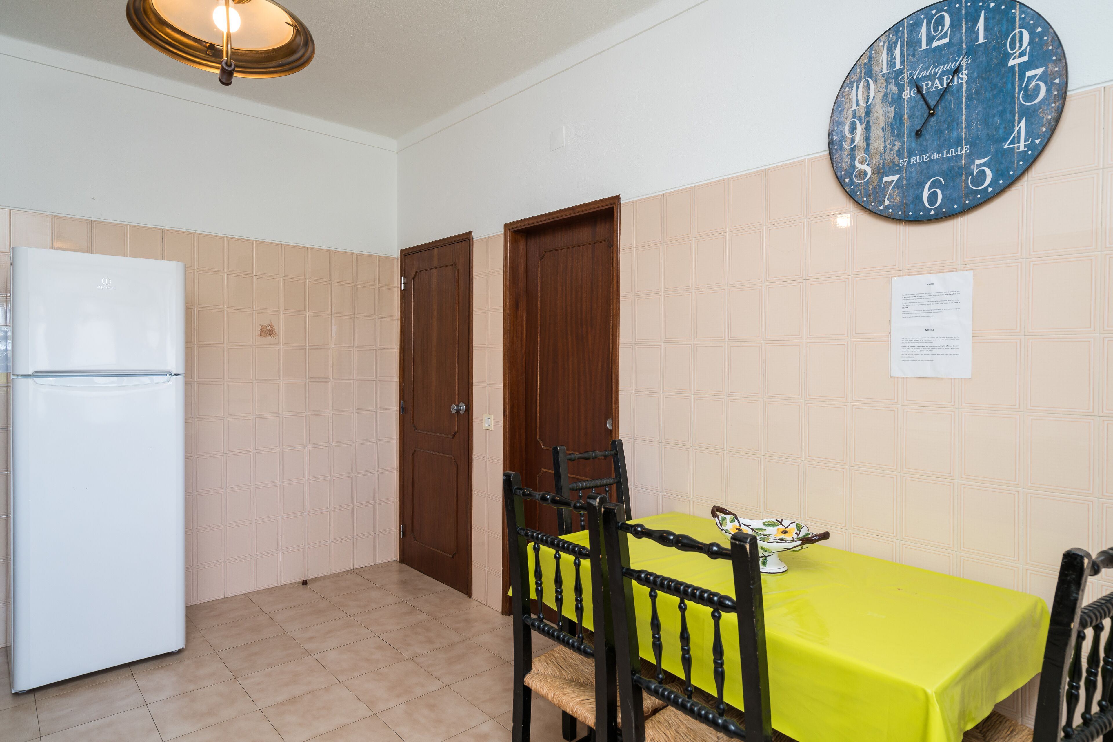 Una cucina con frigorifero, porte in legno, un tavolo da pranzo con tovaglia gialla e una sveglia a parete.