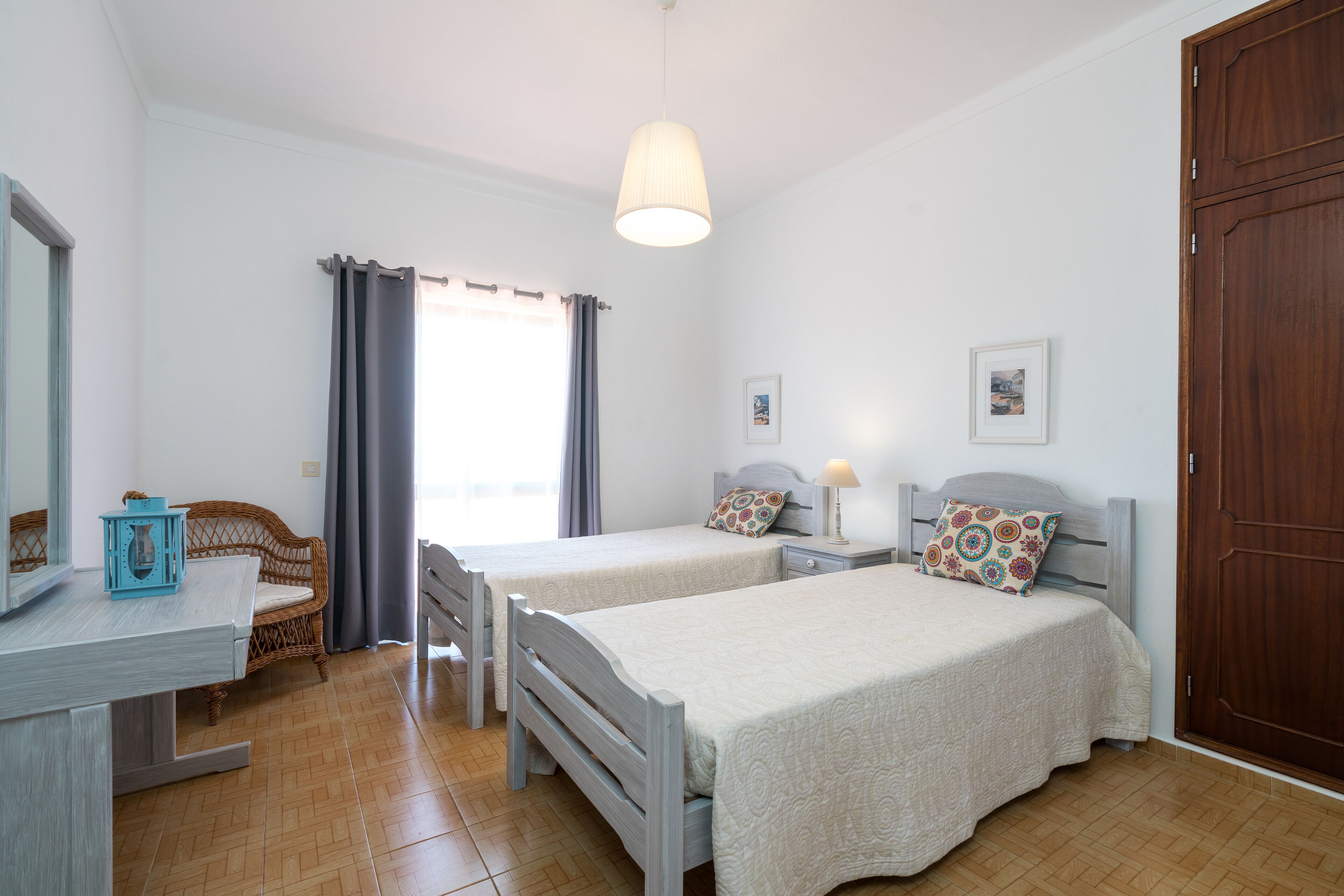 A bedroom with two beds, a wooden wardrobe, a desk with a chair, a window with curtains, and a hanging light fixture.