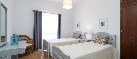 A bedroom with two beds, a wooden wardrobe, a desk with a chair, a window with curtains, and a hanging light fixture.