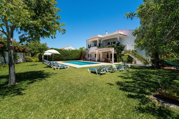 Una casa de dos pisos con piscina, rodeada por un césped cuidado y árboles.