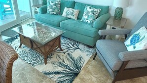 Een woonkamer met een blauwgroene bank, een glazen salontafel, een rieten fauteuil, een kamerkleed met dessin en een bijzettafeltje met lamp.