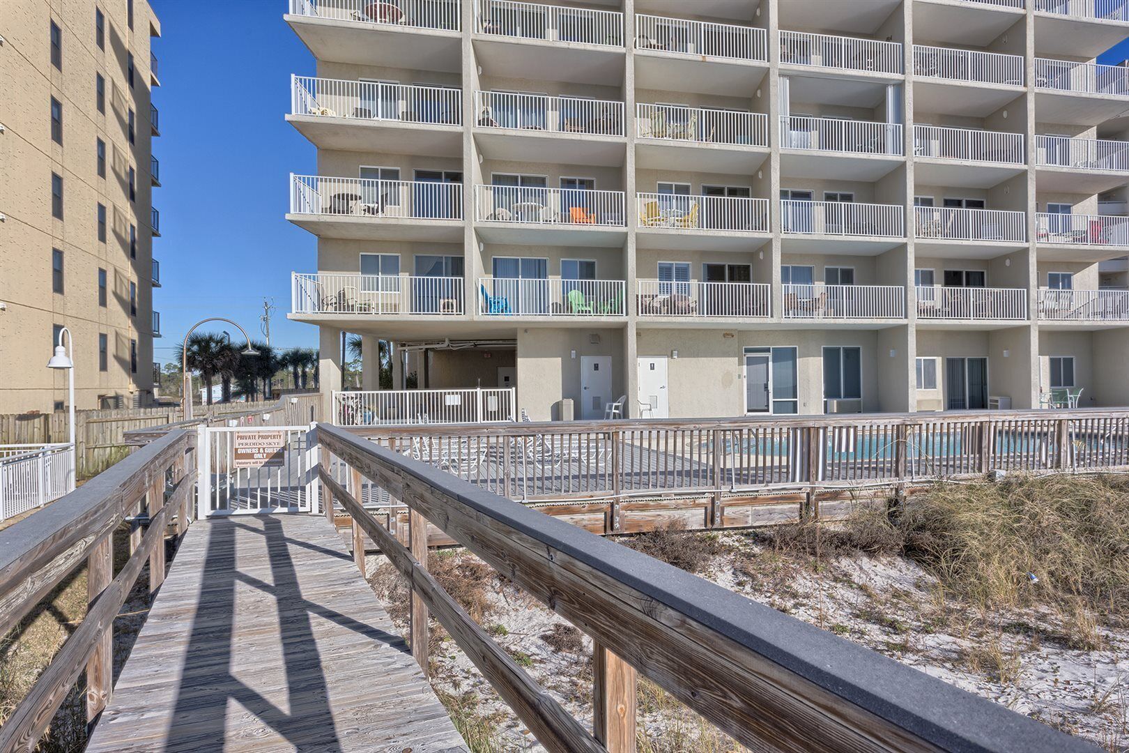 A multi-story building with balconies, a wooden boardwalk leading to a pool, and a sandy beach area.