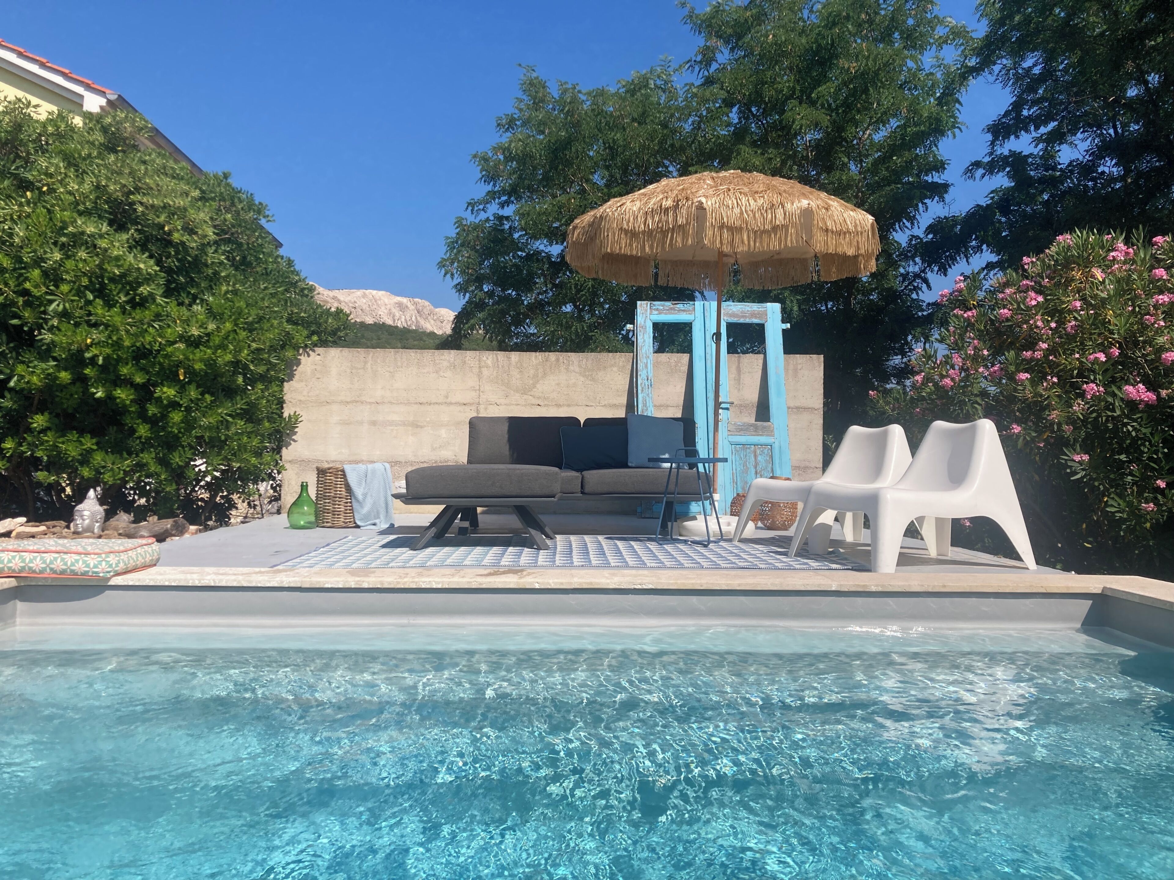 A pool area with a blue-tiled deck, a sofa, two white chairs, a thatched umbrella, and a decorative fountain.