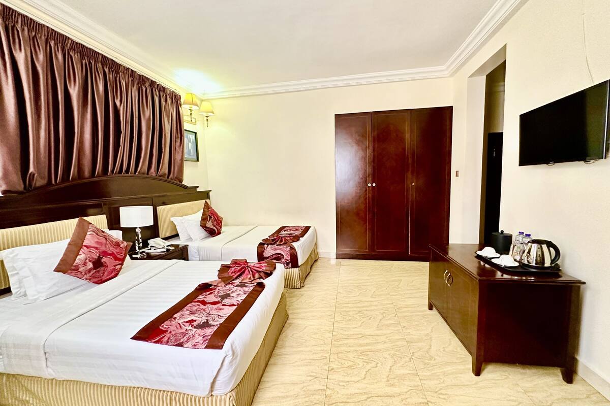 A hotel room with two beds, a wooden wardrobe, a television, and a bedside table with a kettle.