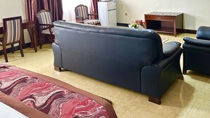 A hotel room with a large sofa, a bed with a patterned bedspread, a television mounted on the wall, and a desk with a computer and a lamp. - Sonia Hotel (Accra)
