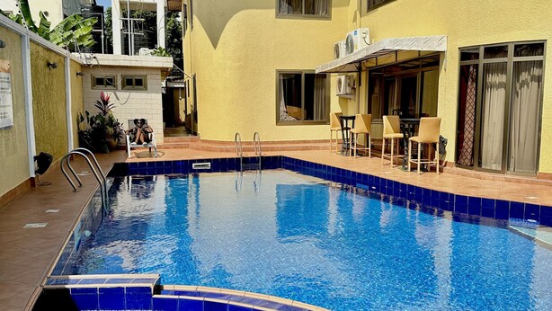 A swimming pool with blue tiles, a sun terrace with chairs, and a building with a yellow exterior. - Sonia Hotel (Accra)