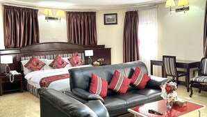 A hotel room with a bed, a black leather sofa, a red coffee table, and a dining area with chairs and a table. - Sonia Hotel (Accra)